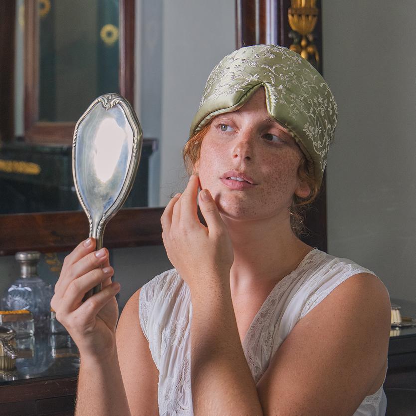 A person wears a sage green patterned Pride & Prejudice - Sage + English Rose sleep mask pushed up onto their forehead while looking into a hand mirror.