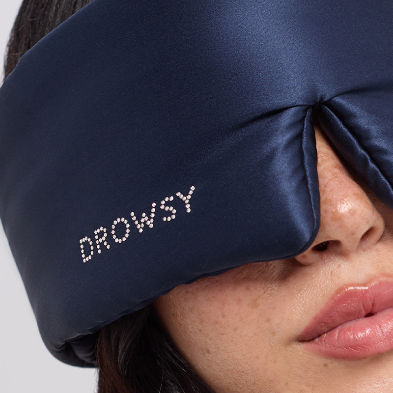A close-up of a person wearing the drowsysleepco Midnight Blue sleep mask, featuring a Swarovski® embellished logo and partially covering their face—the perfect accessory for anyone seeking true beauty sleep.
