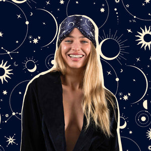 A smiling woman with long blonde hair wears us.drowsysleepco’s Cosmic Energy celestial-patterned silk eye mask and a dark robe, standing before a navy background adorned with moons, suns, and stars.